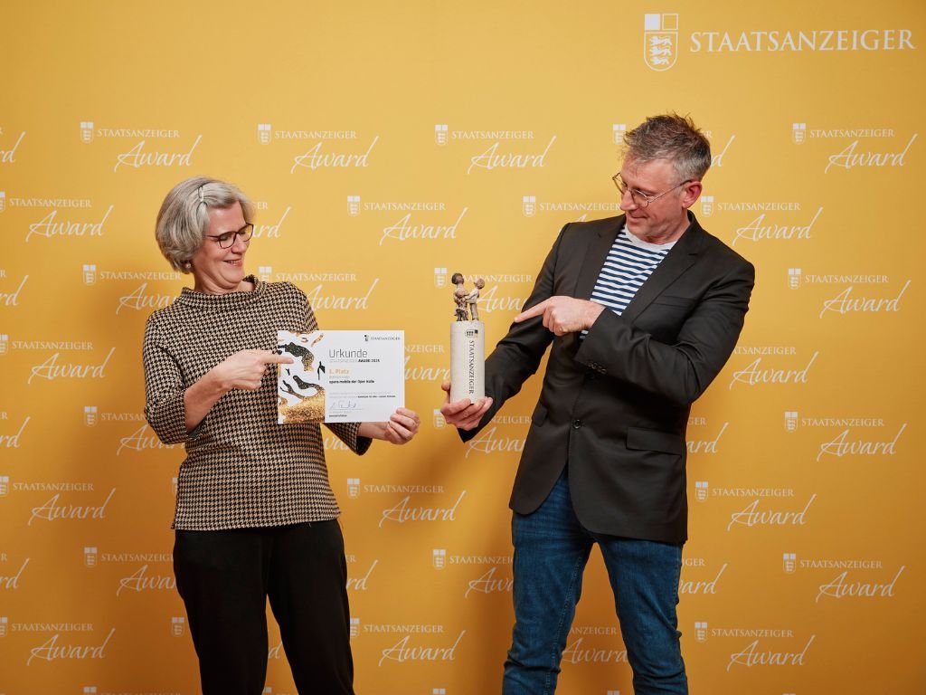 Preisträger präsentieren Urkunde und Trophäe beim Staatsanzeiger Award vor gelbem Hintergrund