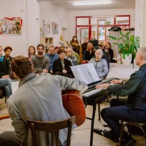 Musiker mit Gitarre und Keyboard treten vor einem gemischten Publikum in einer sozialen Einrichtung auf