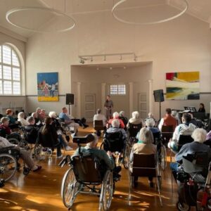 Konzert in einem hellen Saal mit Seniorinnen und Senioren, einige im Rollstuhl, verfolgen die Darbietung