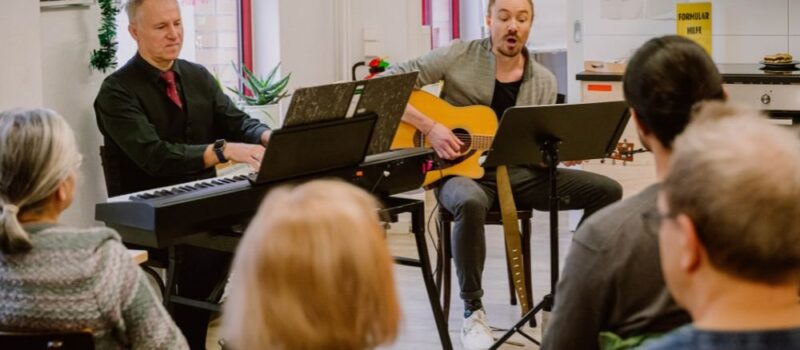 Musiker spielen Gitarre und Keyboard vor sitzendem Publikum in einem Gemeinschaftsraum