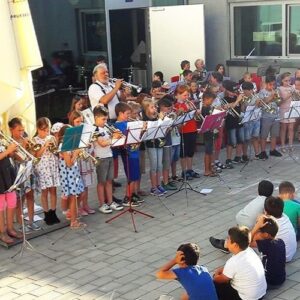Kinderorchester spielt gemeinsam im Freien vor Publikum, begleitet von einem erwachsenen Musiker