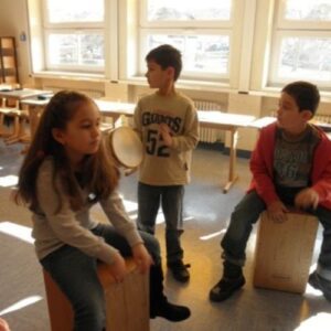 Drei Kinder musizieren im Klassenraum mit Cajón und Tamburin