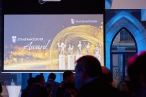 Gäste sitzen im Veranstaltungsraum und blicken auf eine Präsentationsfolie zum Staatsanzeiger Award mit dargestellten Trophäen auf einer großen Leinwand.