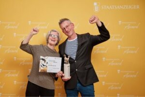 Zwei Preisträger heben jubelnd die Arme und präsentieren Urkunde und Trophäe beim Staatsanzeiger Award 2025.