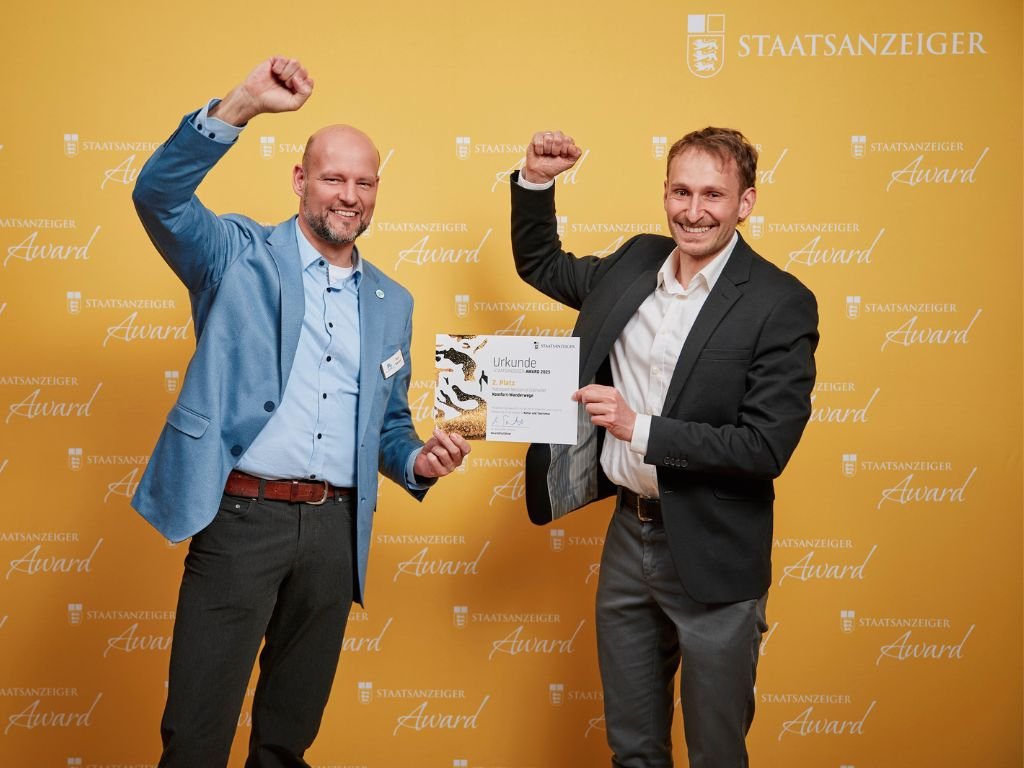 Zwei Männer präsentieren Urkunde beim Staatsanzeiger Award vor gelbem Hintergrund