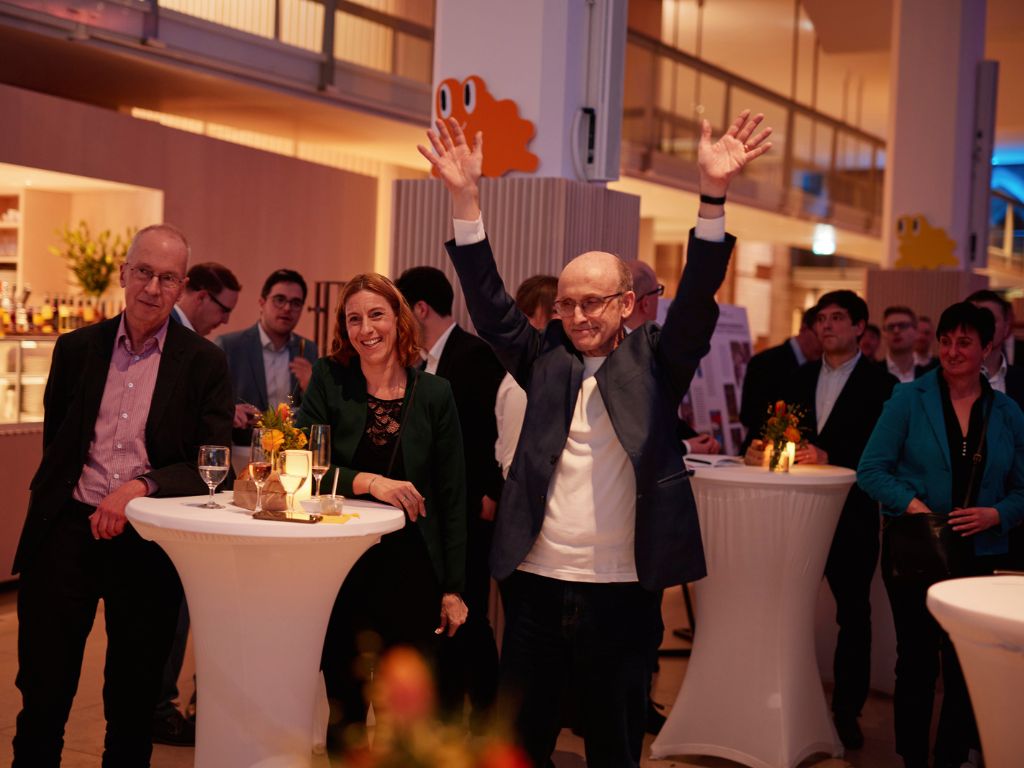 Teilnehmende einer Veranstaltung stehen an Stehtischen in einem Innenraum; ein Mann hebt die Arme, während andere Gäste bei einem Empfang miteinander ins Gespräch kommen.