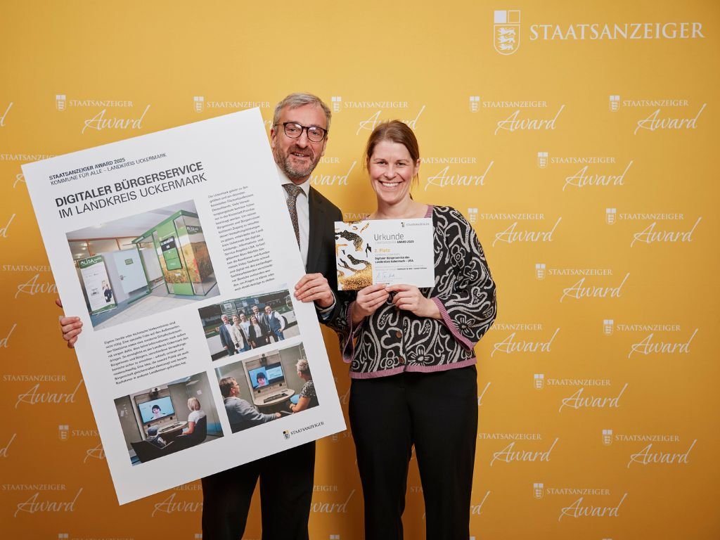 Zwei Personen halten Urkunde und Präsentationsposter zum digitalen Bürgerservice Uckermark vor gelbem Award-Hintergrund